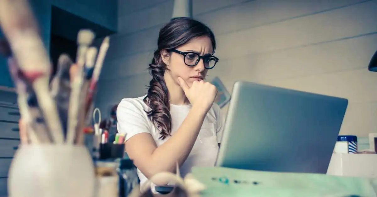 Junge Frau sitzt an einem Tisch vor einem Laptop und denkt angestrengt nach. Der Tisch ist etwas unordentlich. Die Frau trägt Brille, ein weisses Shirt und hat die Haare zu einem Zopf gebunden.