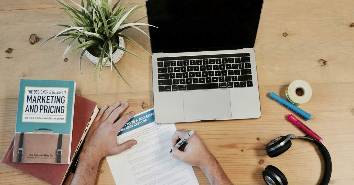 Tisch mit Laptop und verschiedenen Arbeitsutensilien. Eine Hand die auf ein Blatt Papier schreibt.